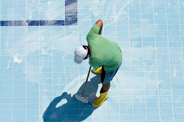 Pool technician cleaning thick mineral deposits from raised spa tile beside a backyard pool, Experienced pool cleaner inspecting hard water scale on decorative waterline tile